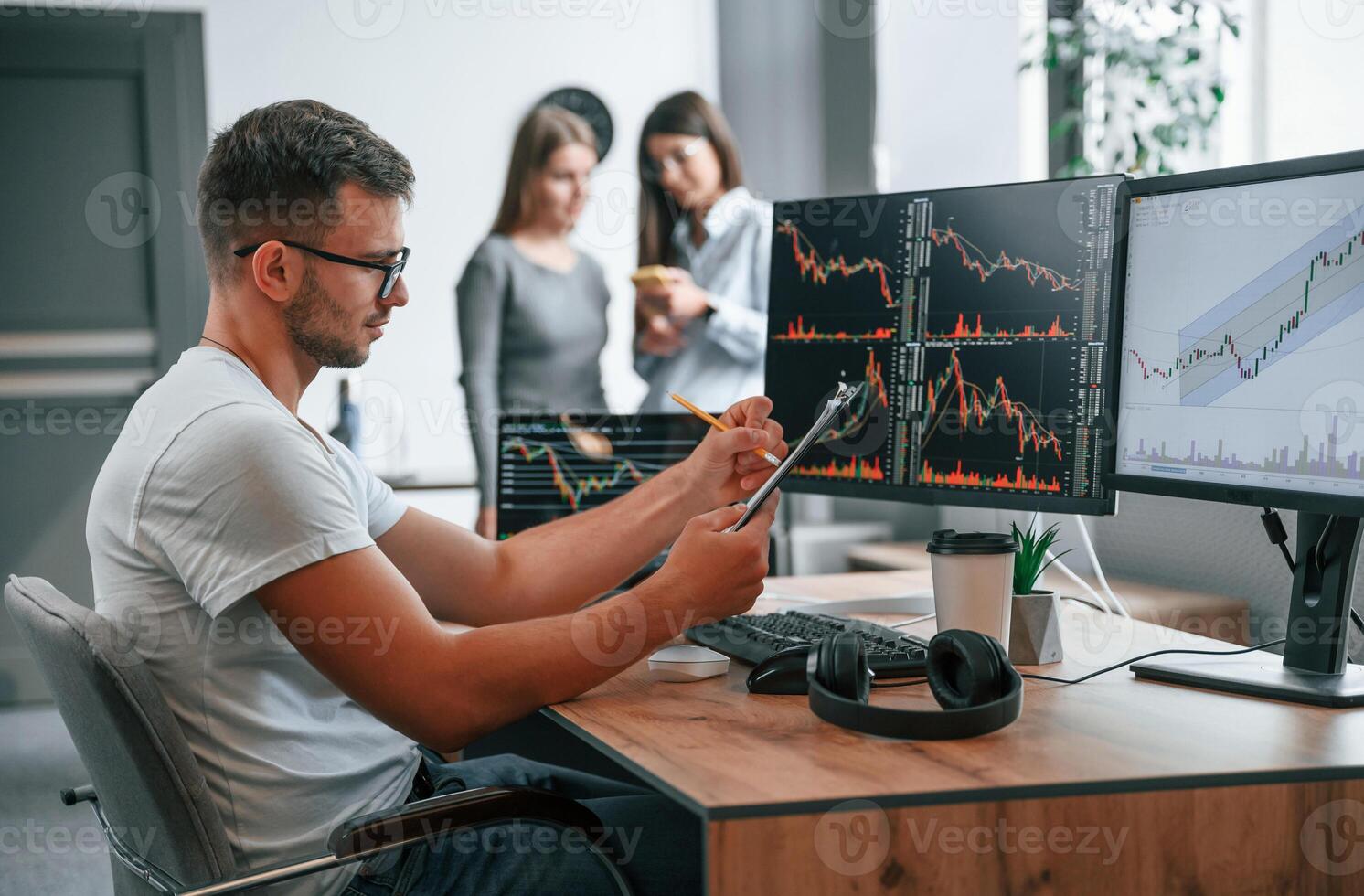 Stock, broker is sitting by computer and working with exchange technology. Women is standing and talking behind photo