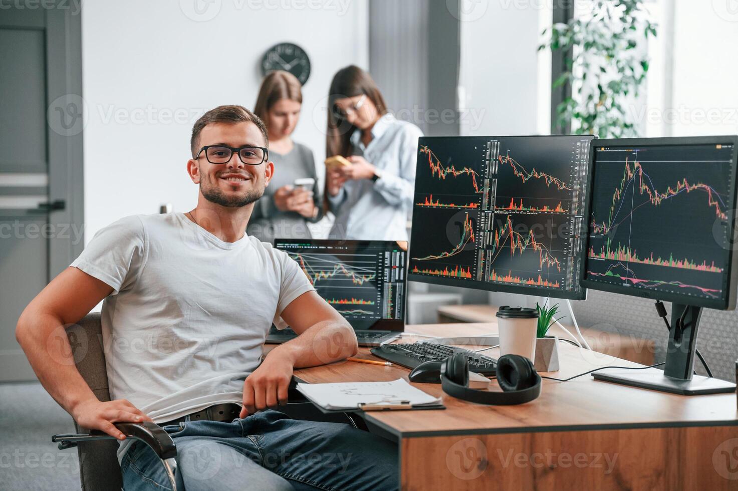 Stock broker, is sitting by computer and working with exchange technology. Women is standing and talking behind photo