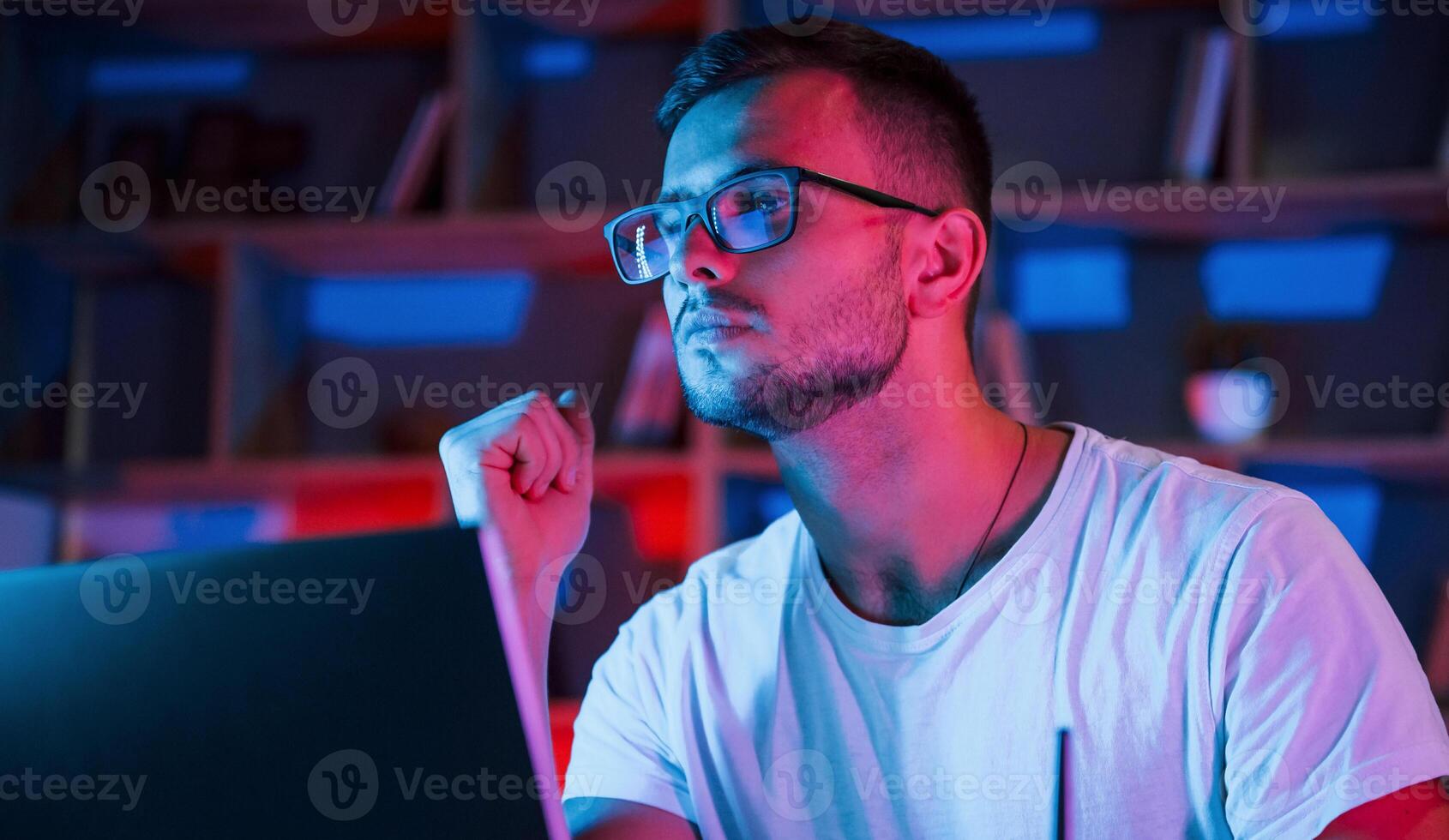 Man in glasses and, white shirt is sitting by the laptop in dark room with neon lighting photo