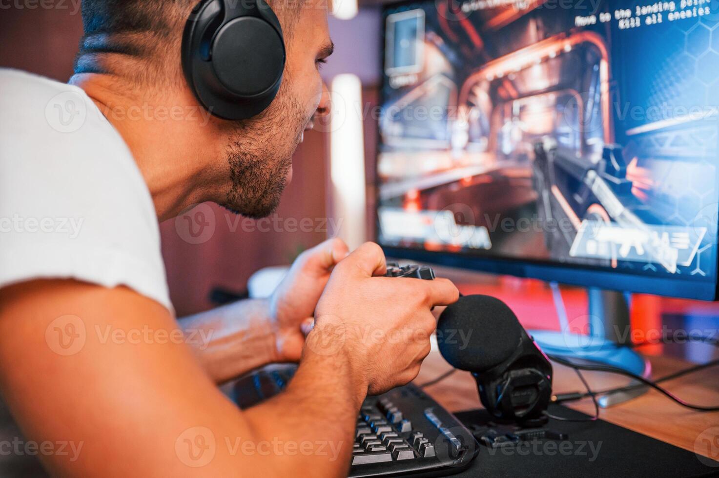 Playing first, person shooting game. Male streamer in casual clothes is indoors with pc photo