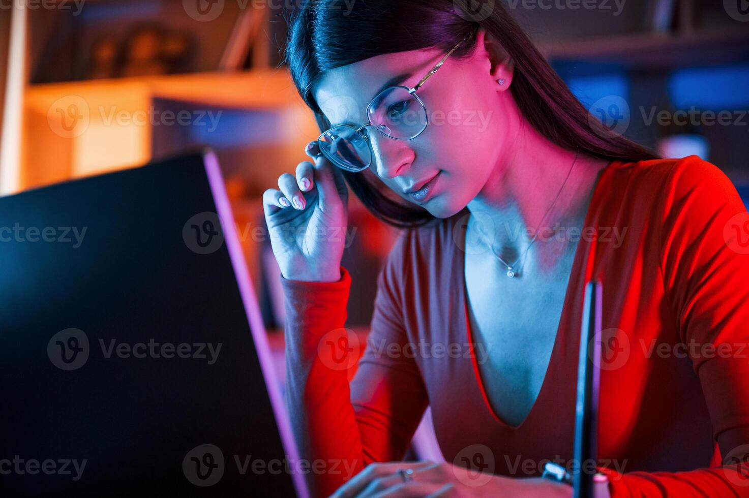 Focused on the work.. Beautiful woman in glasses and red wear is sitting by the laptop in dark room with neon lighting photo