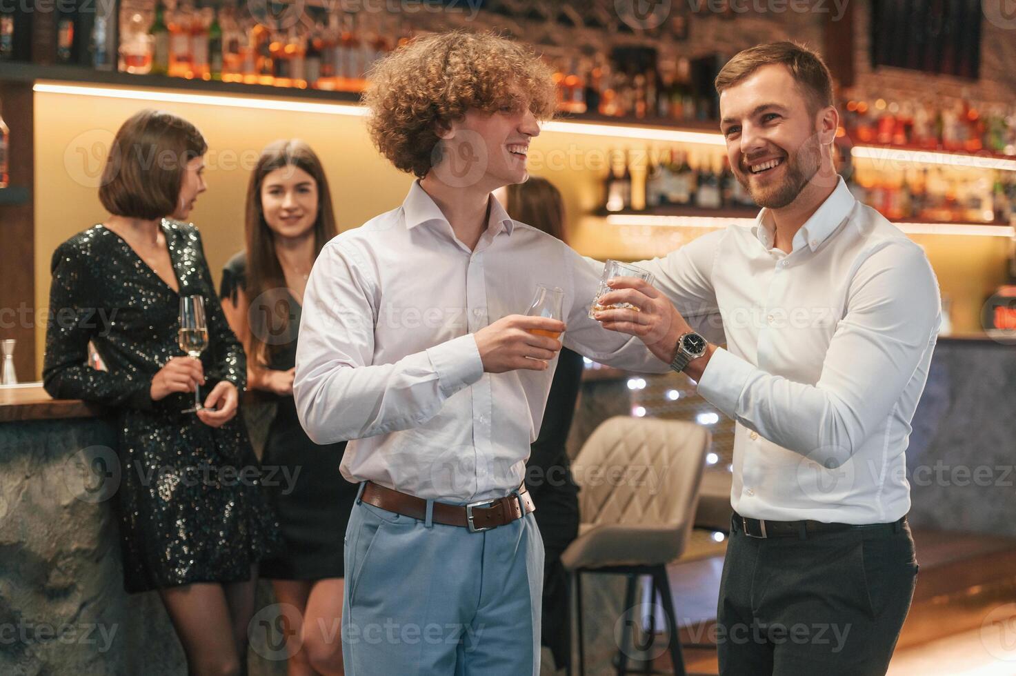 Two men, are standing together in formal clothes on the party event. Friends are behind photo