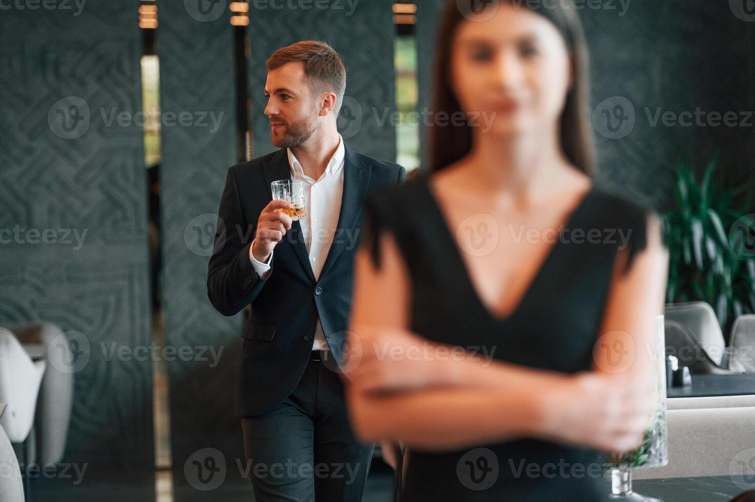 Man in black suit and ,with drink in hands looks at woman in dress photo