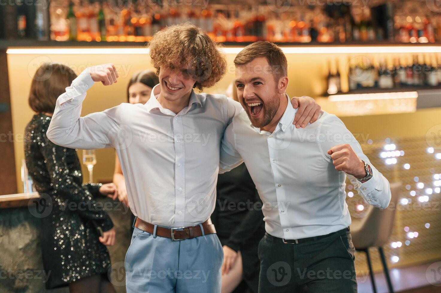 Two men are standing together in formal ,clothes on the party event. Friends are behind photo