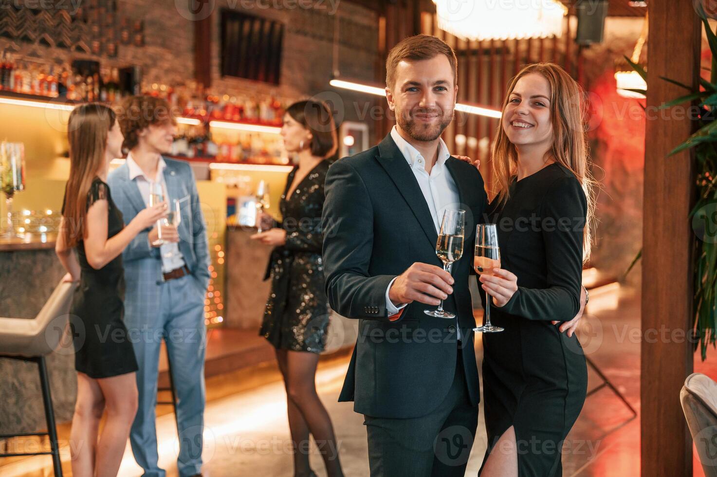Man with his woman is standing and smiling. Group of people in beautiful elegant clothes are celebrating New Year indoors together photo