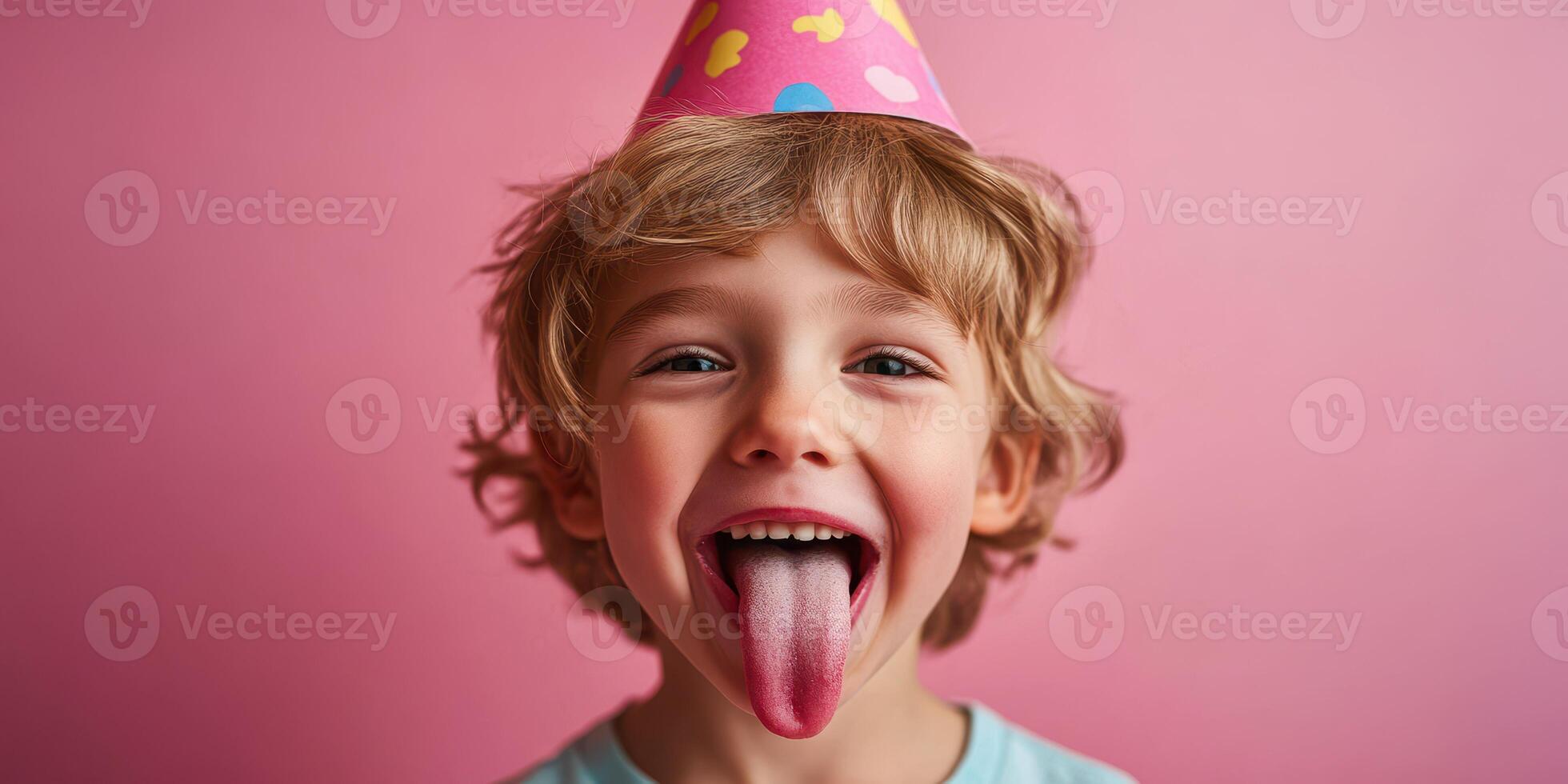 Child celebrates birthday with party hat, playful expression, and sticky tongue against a pink background photo