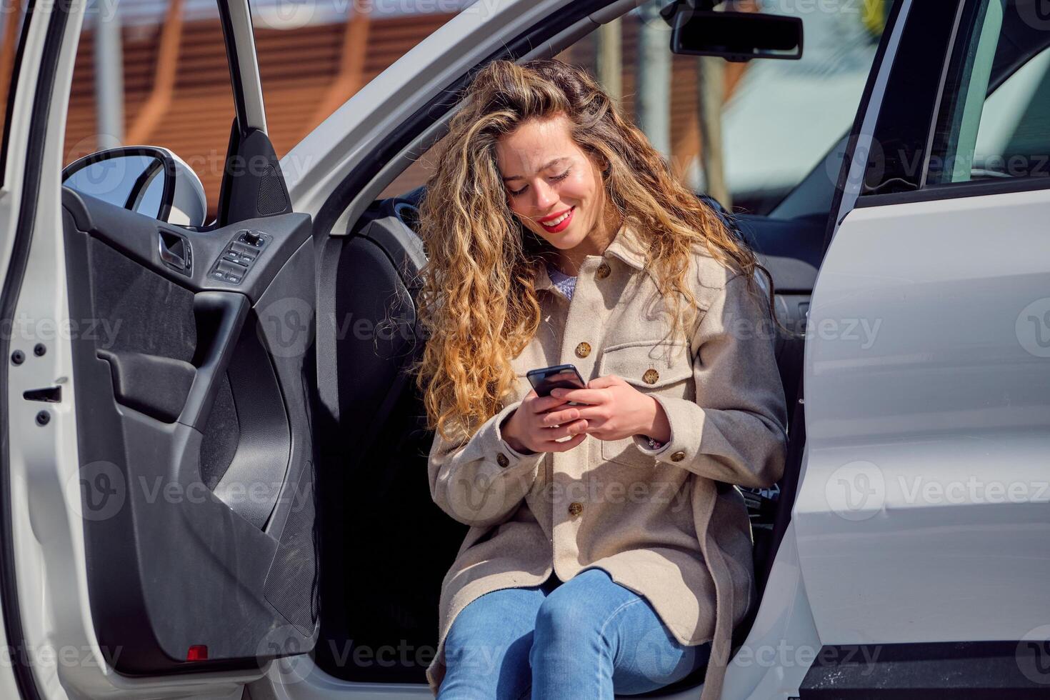 Smiling woman in coat browsing smartphone and sitting in car photo