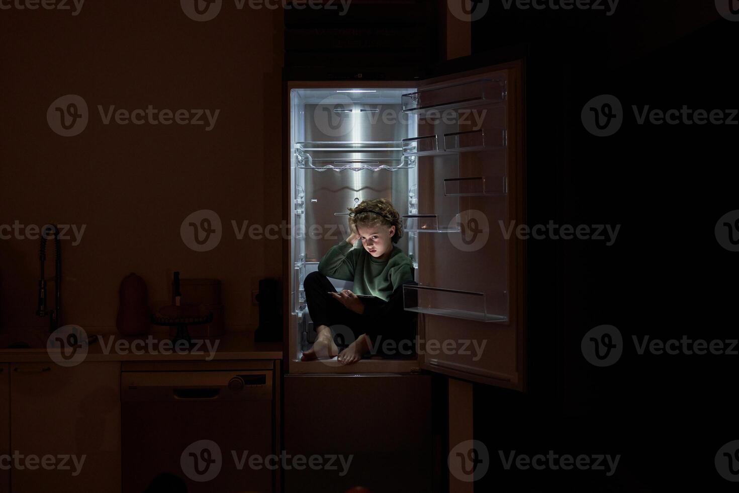 Thoughtful boy with tablet sitting in fridge at night photo