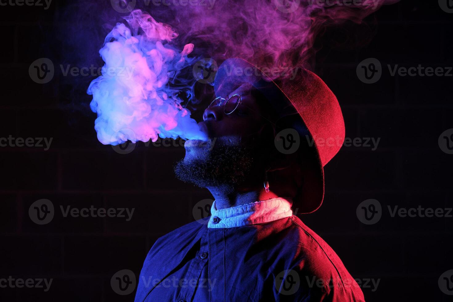 Stylish black man in hat vaping against brick wall photo