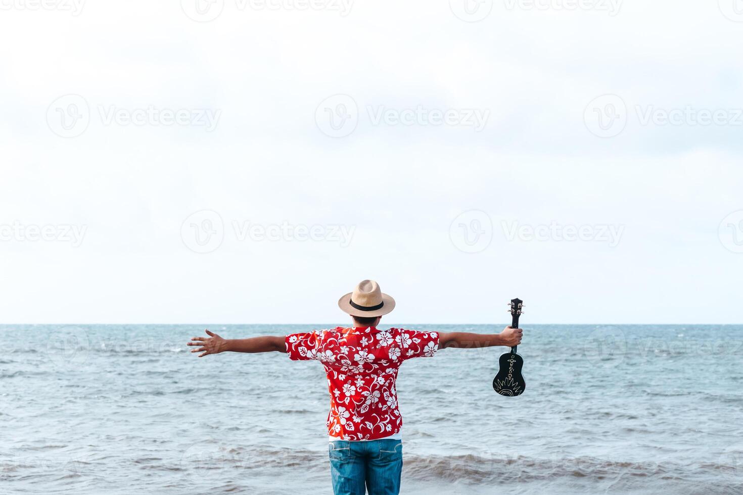 A man with colorful hat and a ukulele outfit holding his hands in the air in front of the sea. freedom concept. photo