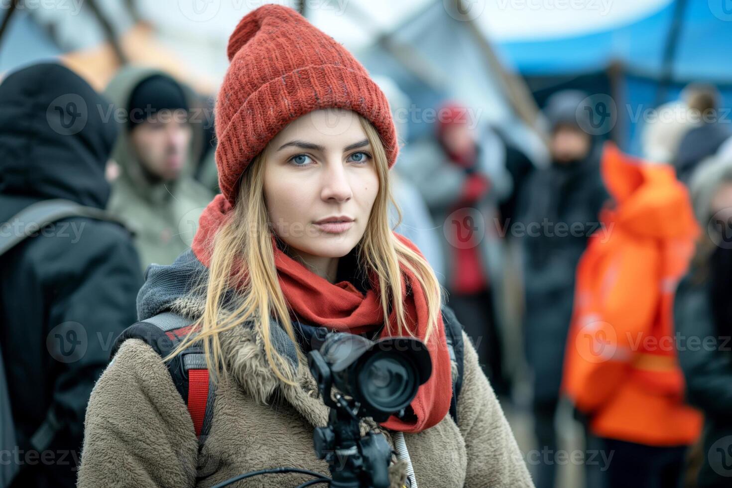 A young Caucasian woman with long blonde hair wearing a cozy brown coat and red beanie, holding a camera at an outdoor event. photo