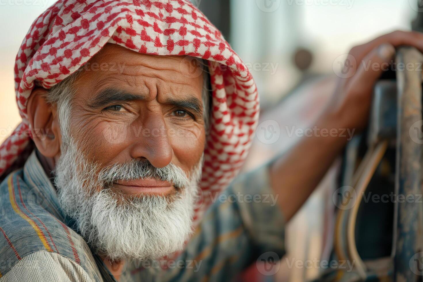 An older Middle Eastern man with a white beard and red checkered keffiyeh looks thoughtfully at the camera, showcasing his life experience. photo