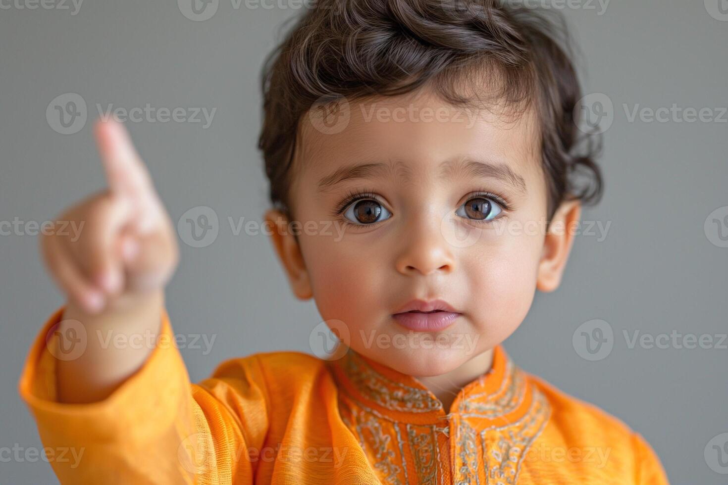 A young boy of South Asian descent, wearing an orange traditional outfit, curiously gestures with his finger while looking at the camera. photo