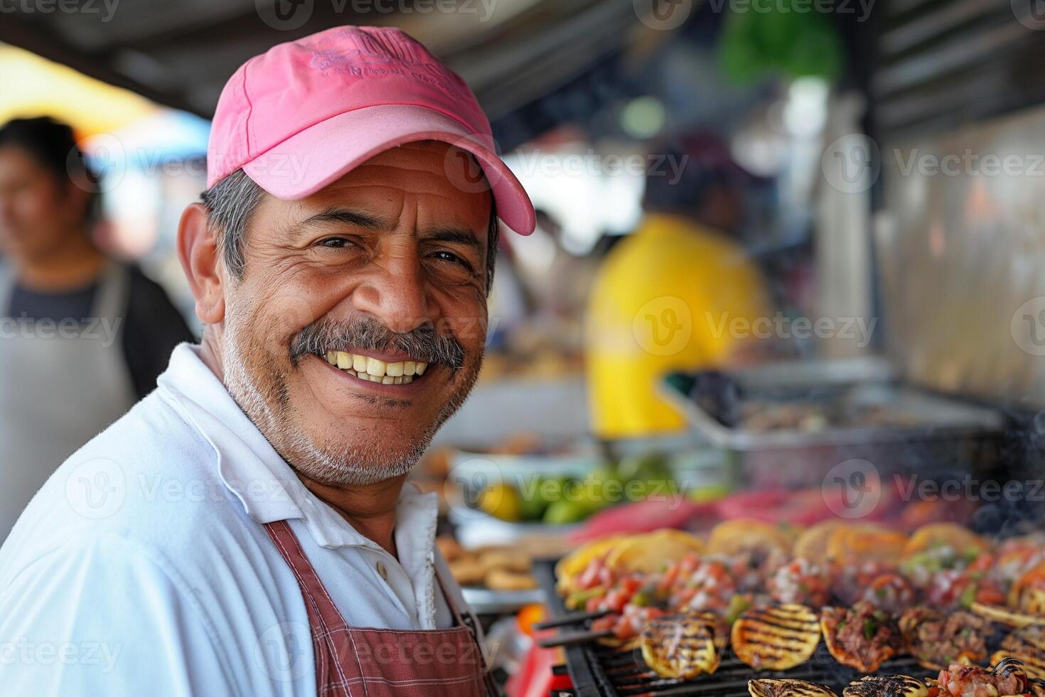 A smiling Hispanic man in a pink cap stands by a grill, showcasing delicious grilled food at a vibrant outdoor market. photo