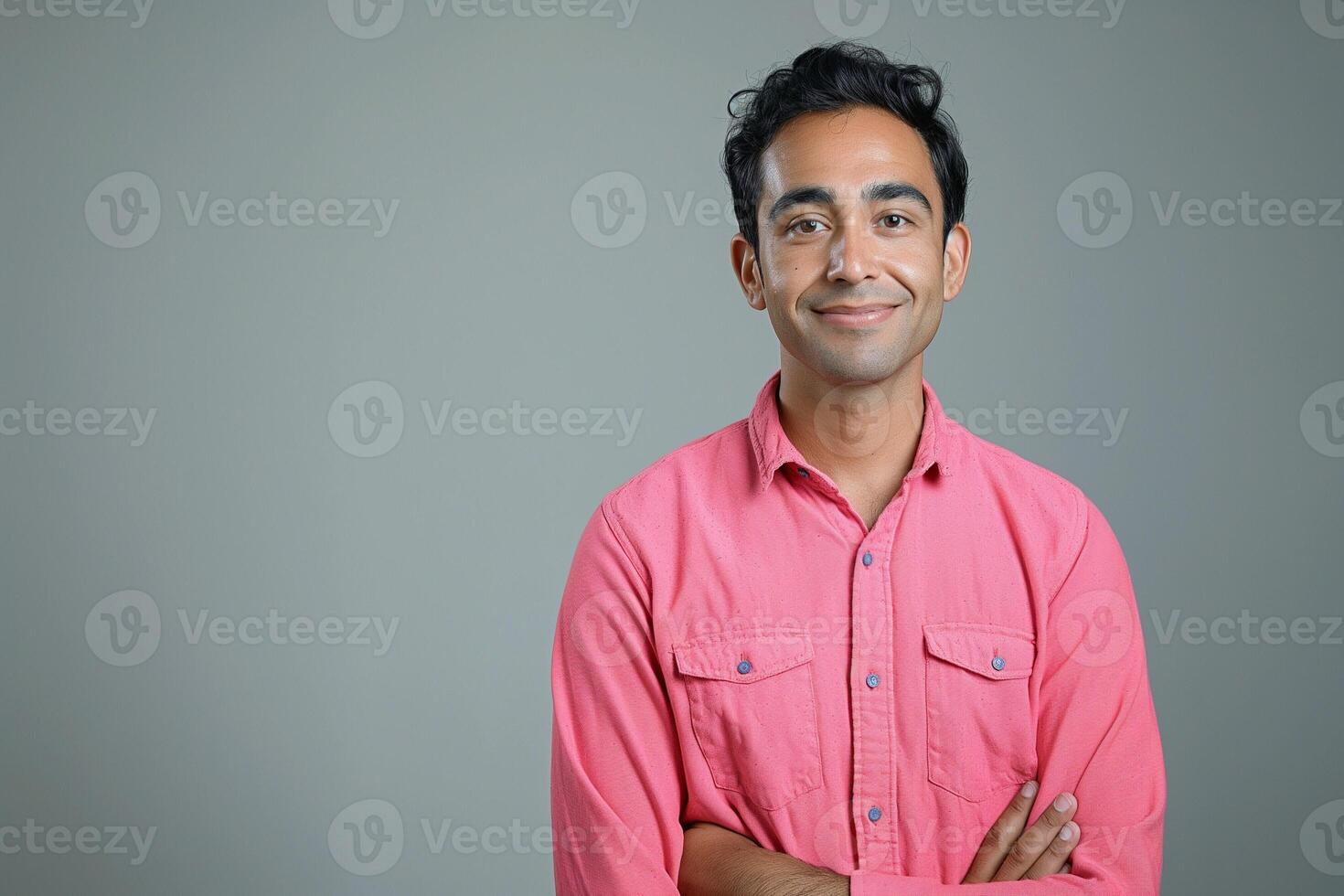 Smiling South Asian man wearing a pink button-up shirt, arms crossed, against a neutral gray background. photo