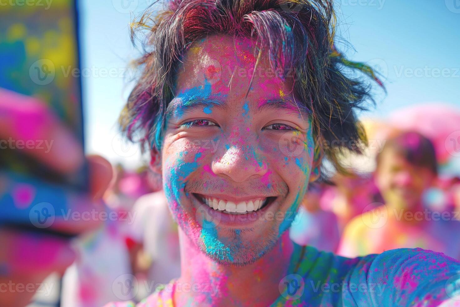 A cheerful Asian male celebrating Holi, covered in vibrant colors, captures a selfie amid a festive atmosphere. photo