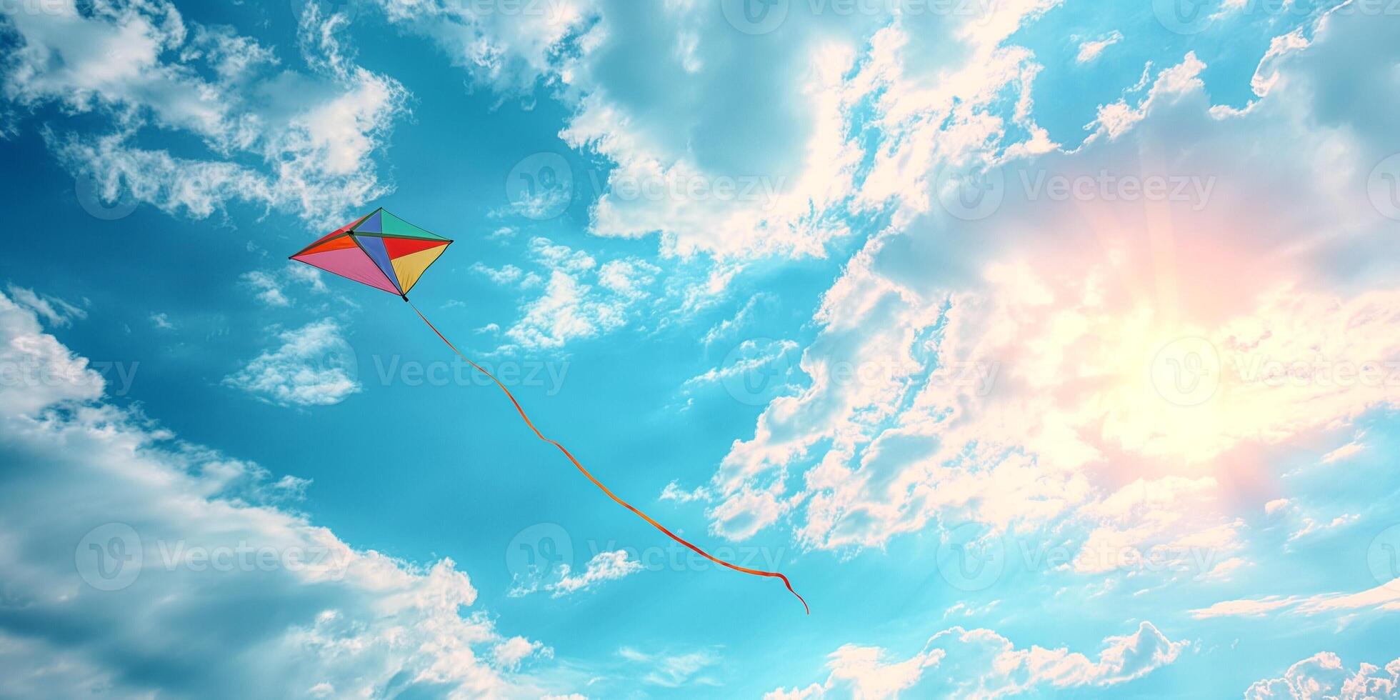 A colorful kite soars high against a vibrant blue sky with fluffy clouds and sunlight streaming through. photo