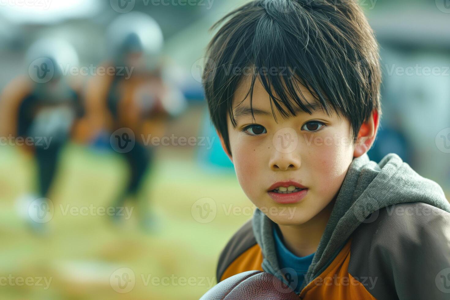 A focused young Asian boy holding a football during practice, showcasing determination amidst teammates training in the background. photo