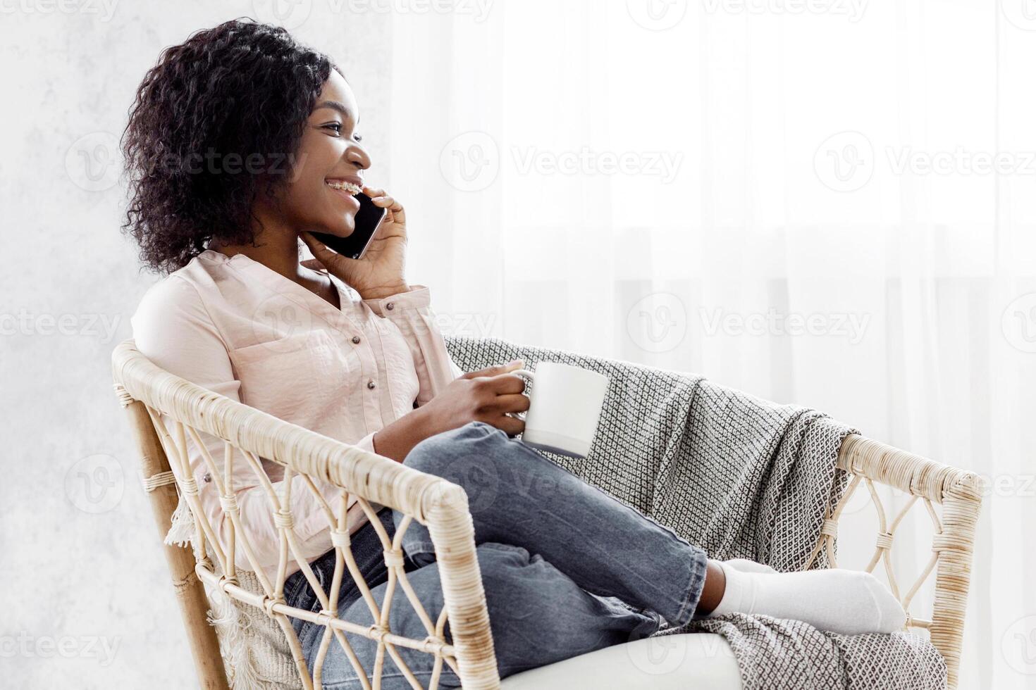 Happy Black Woman Relaxing In Wicker Armchair, Drinking Tea And Talking On Cellphone, Side View With Empy Space photo