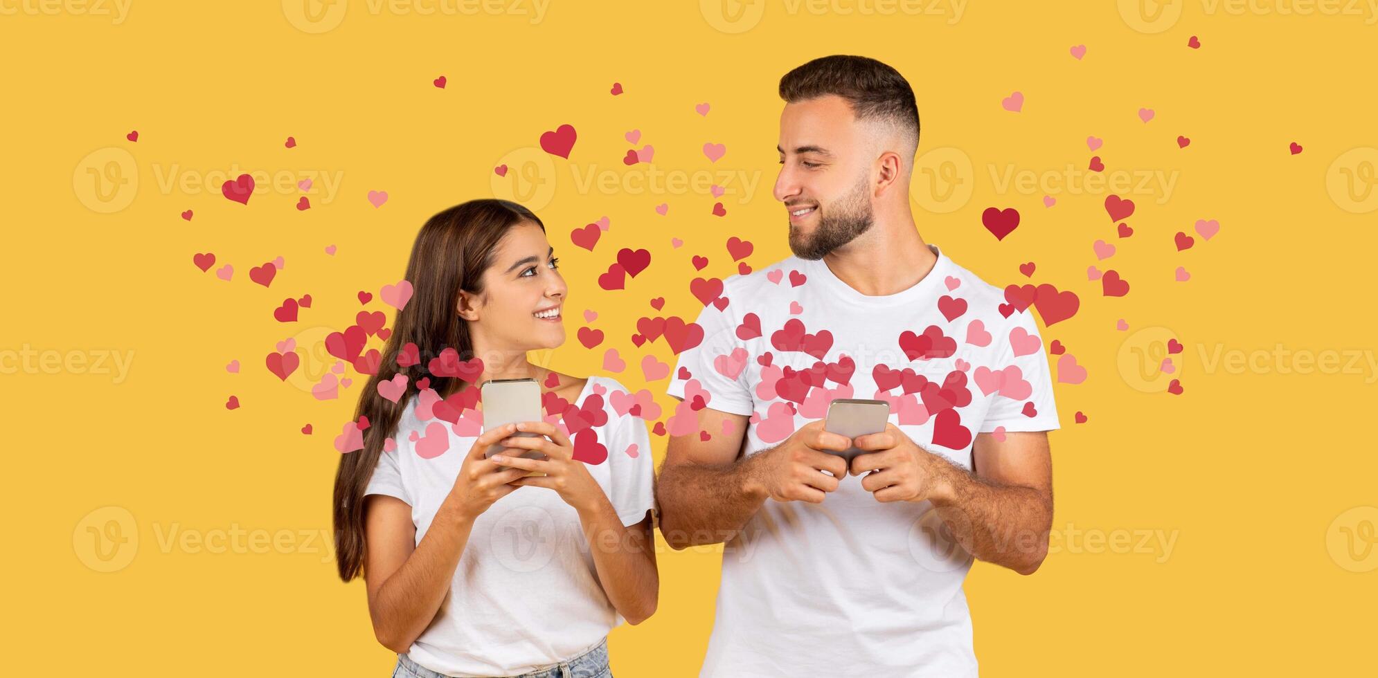 A couple shares a joyful moment on Valentine's Day, smiling at each other while holding smartphones. Colorful hearts surround them, adding to the affectionate atmosphere of their celebration. photo