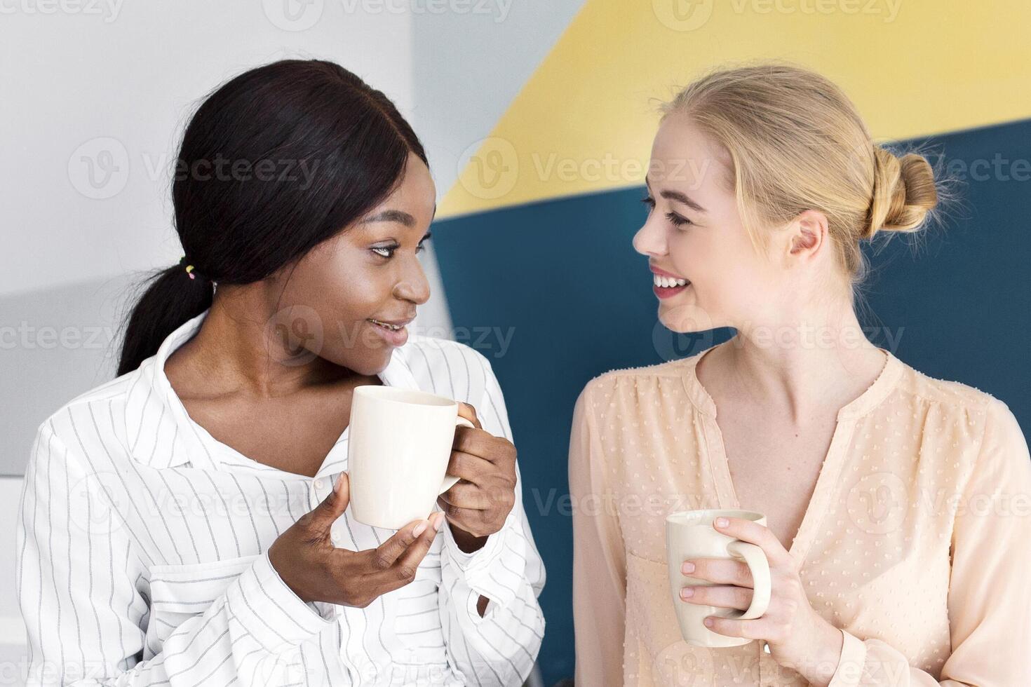 Female call centre operators having coffee and talking during their break at modern open space office, copy space. Positive hotline consultants having friendly chat, enjoying each other's company photo