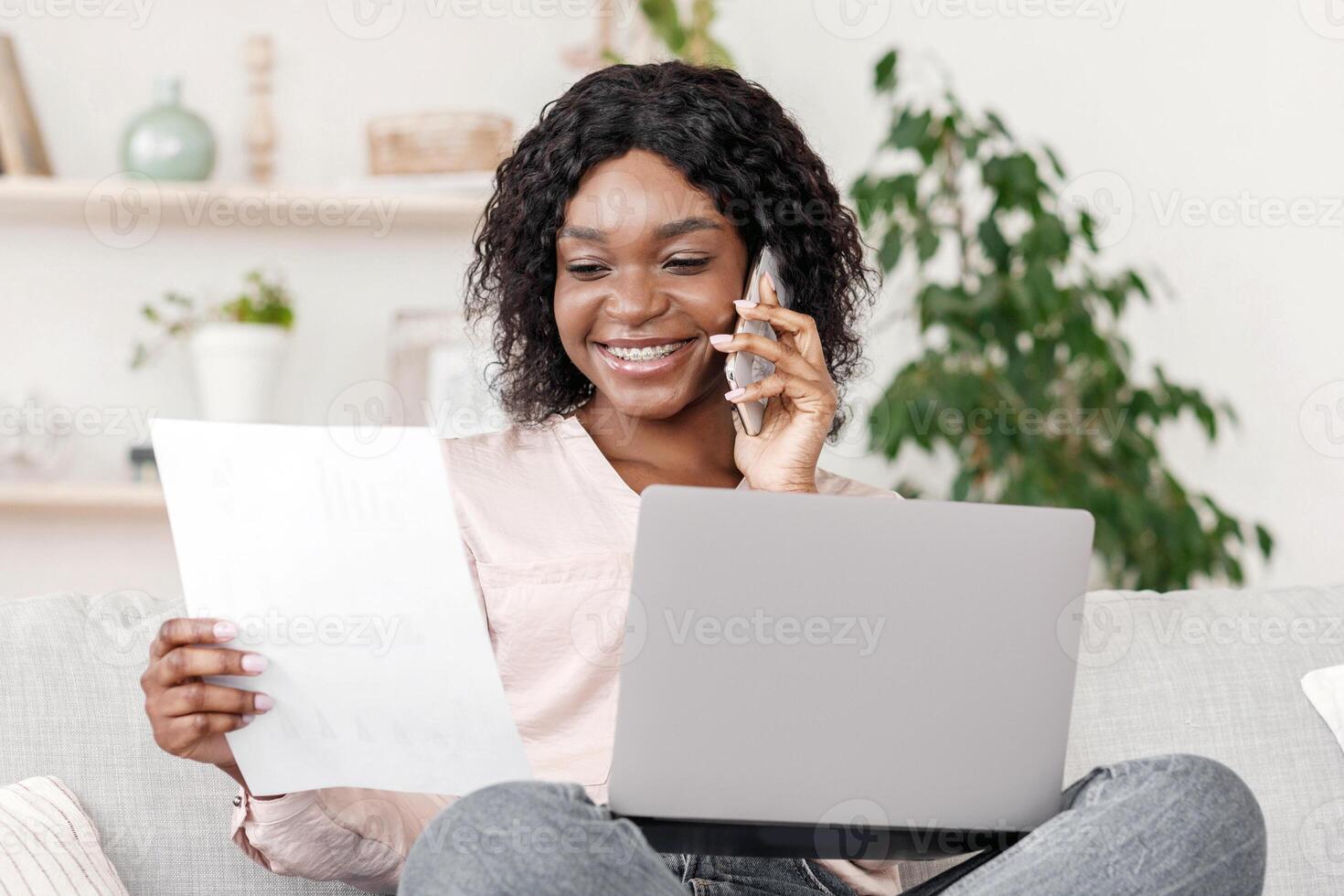 Young black female entrepreneur working from home, discussing documents with client on cellphone, free space photo