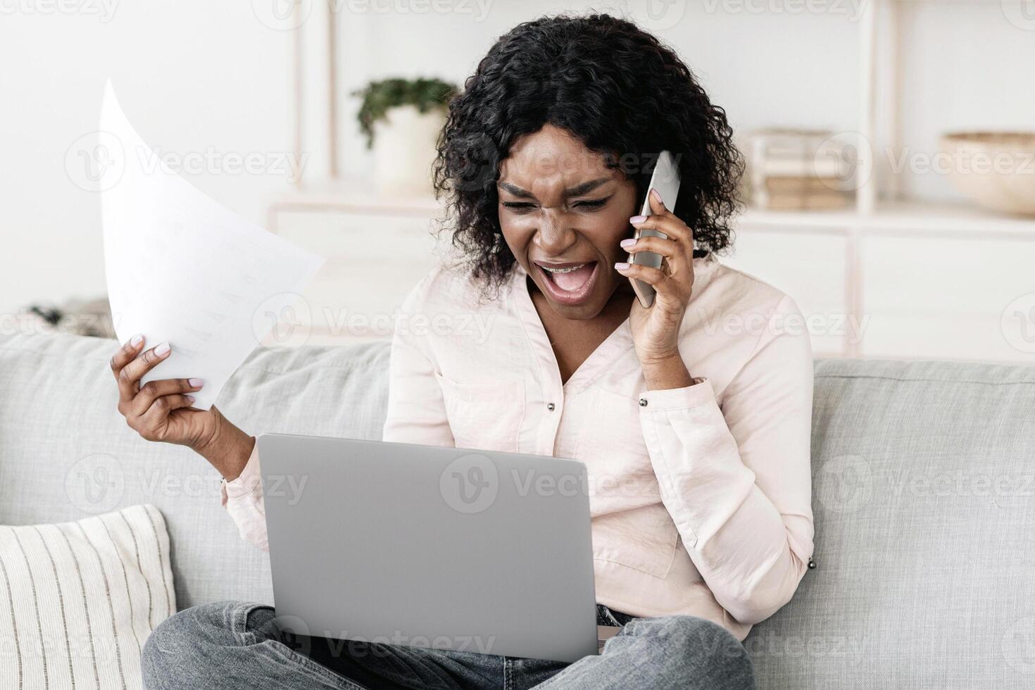 Furious black woman talking on cellphone while working with papers and laptop at home, having business problems, arguing with somebody by phone photo