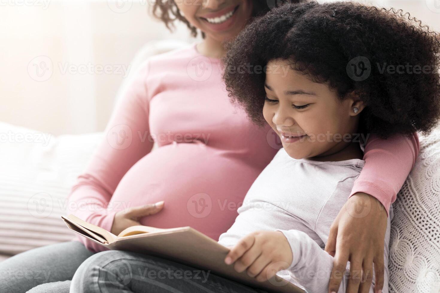 Loving Pregnant Mom And Her Cute Little Afro Daughter Reading Interesting Book Together, Sitting On Couch, Resting At Home, Selective Focus photo