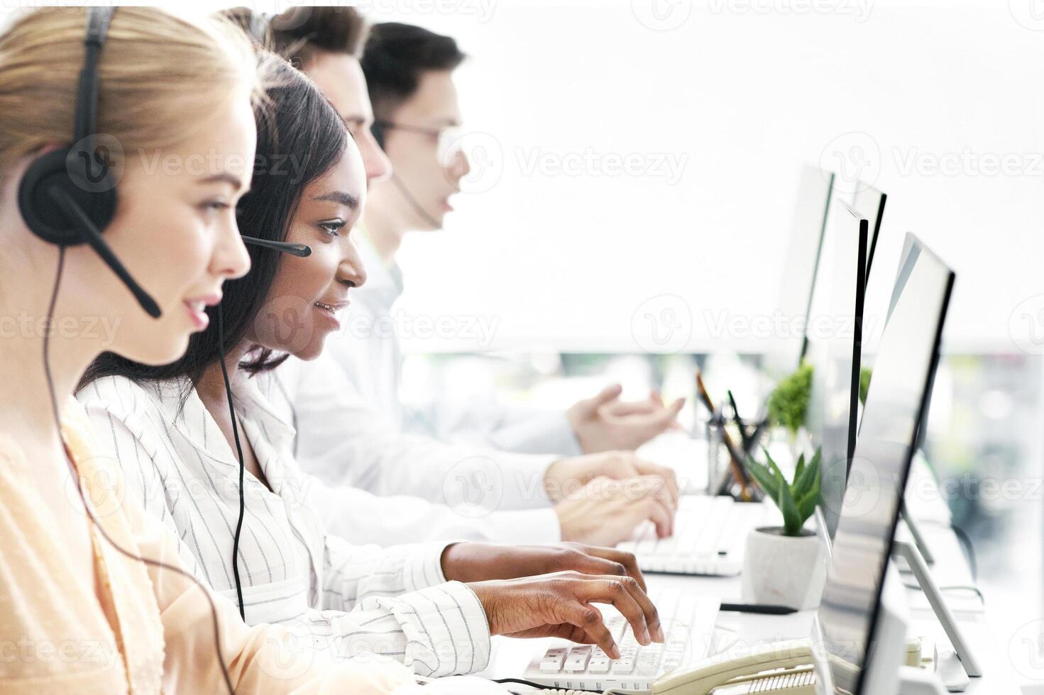Side view of helpline operators with headsets consulting customers at modern call center, empty space. Telemarketing or tech support team working with their computers at open space office photo