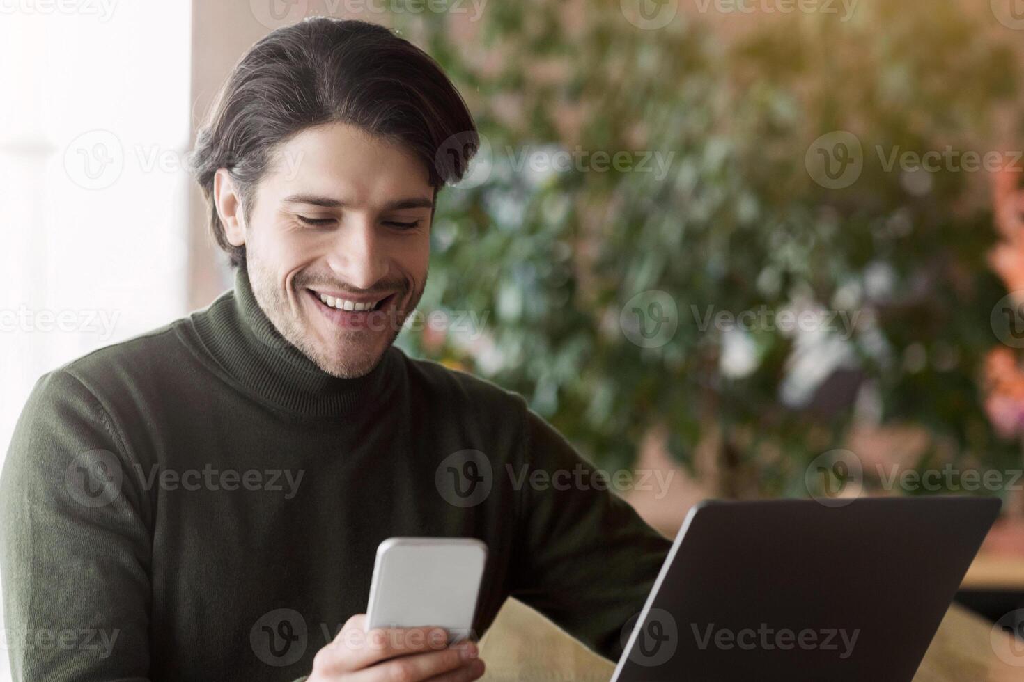 Wealthy young businessman using phone while working with laptop at cafe, time for entertainment, copy space photo