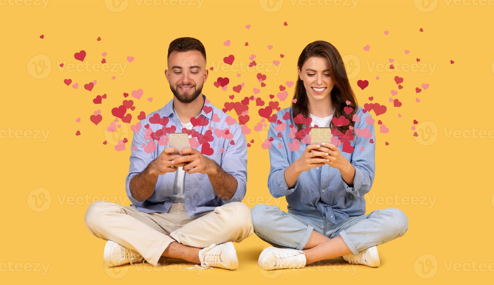 A joyful couple sits together on a yellow background, each holding a smartphone while smiling. They express affection and connection through online messages filled with hearts, celebrating St. photo