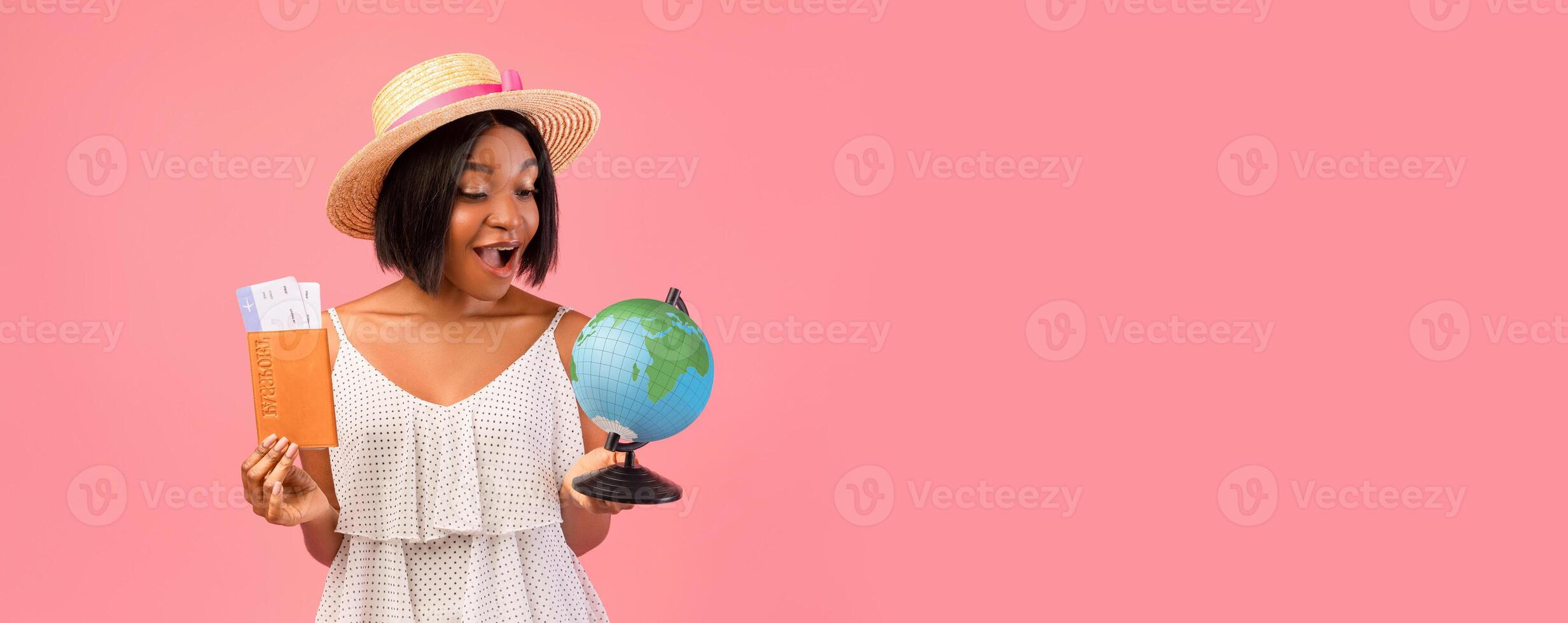Happy African American woman in summer clothes holding globe, passport and plane tickets, traveling around world, having abroad trip during summertime vacation over pink studio background photo