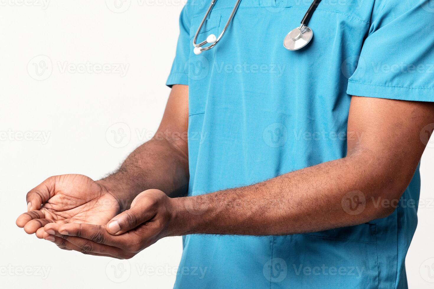 A millennial African American man doctor in scrubs extends his hands with a caring expression. He represents healthcare and patient support in a clinical environment, cropped photo