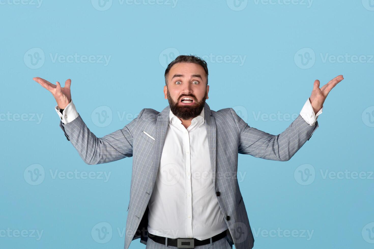 Emotional millennial office worker shrugging his shoulders against blue studio background. Young businessman expressing uncertainty or doubt, feeling confused, having no idea what to do photo