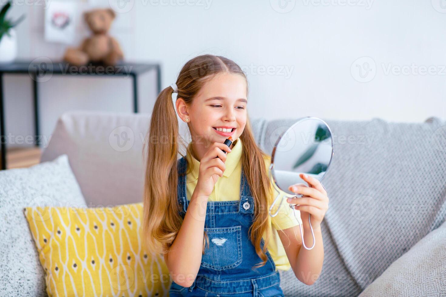 Everyday Make Up. Portrait of cheerful teenage lady applying lipstick, holding mirror and looking at herself with smile, sitting on the couch at home, getting ready for school or date photo