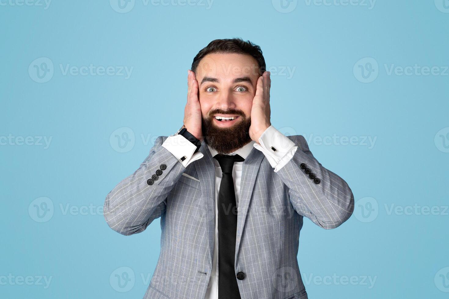 Portrait of shocked young businessman holding his head in excitement, cannot believe his luck or success on blue studio background. Positive millennial CEO feeling astonished and amazed photo