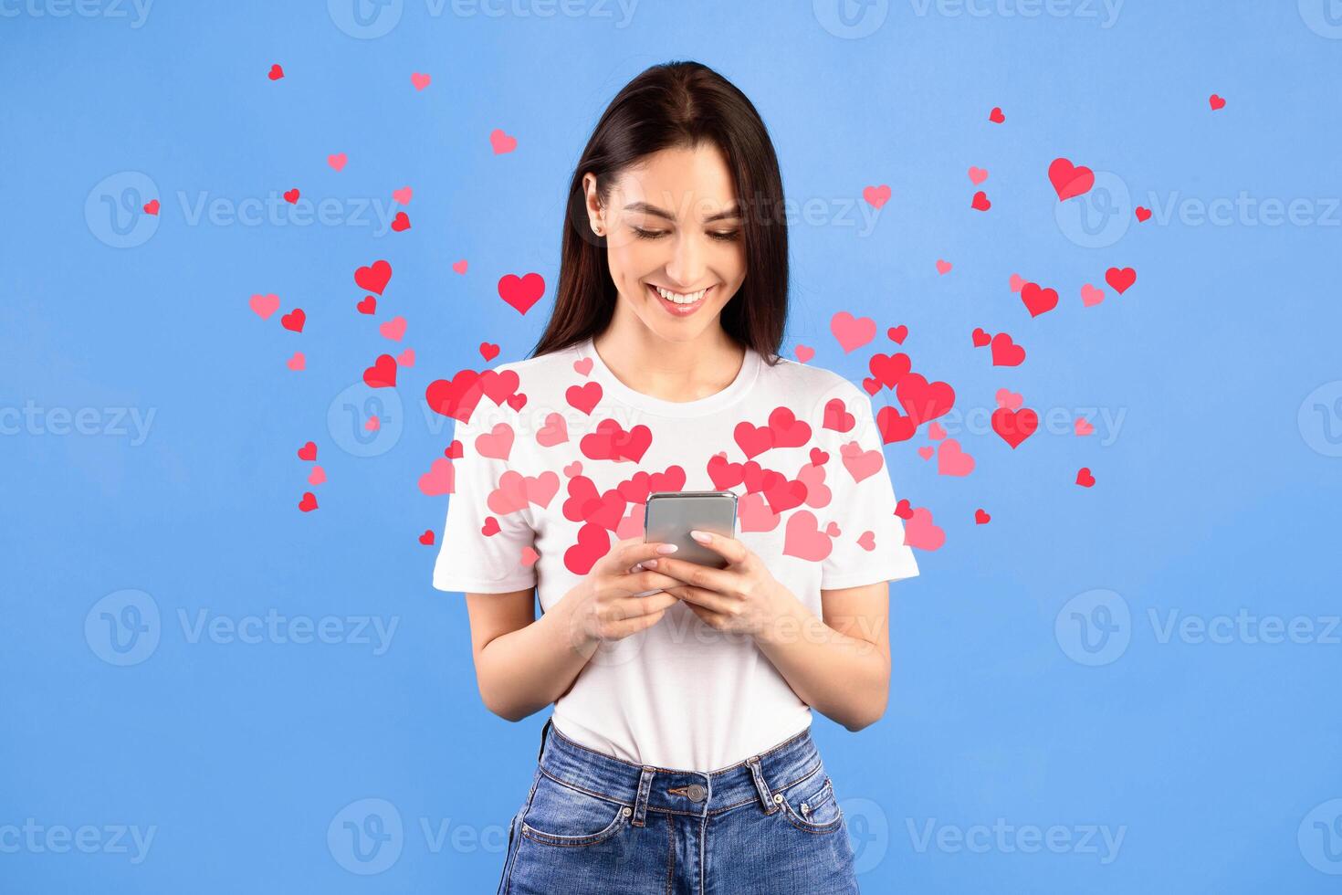 Beautiful happy woman sending love text message on mobile phone with pink hearts flying away from screen isolated on blue studio background. Positive human emotions face expressions photo