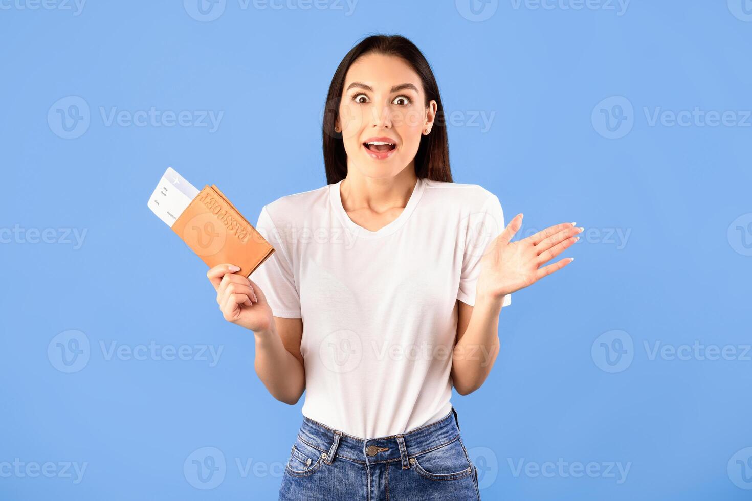 Cheap Flights Concept. Portrait of surprised shocked woman holding passport and boarding pass ticket, standing isolated on blue studio background. Excited lady going on vacation after lockdown photo