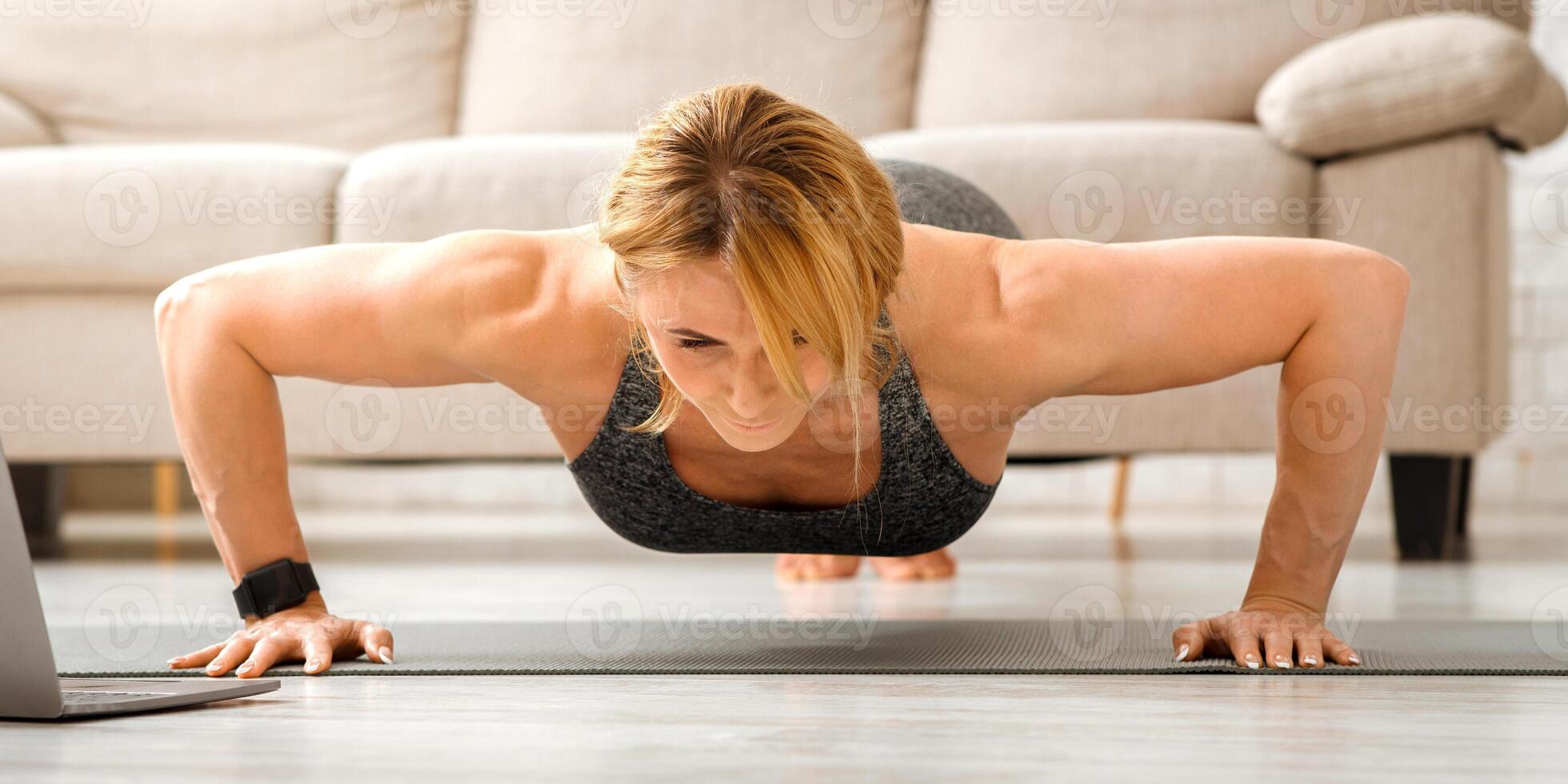 Push ups at home. Adult woman in sportswear doing fitness exercises on floor mat with laptop in living room interior, copy space photo