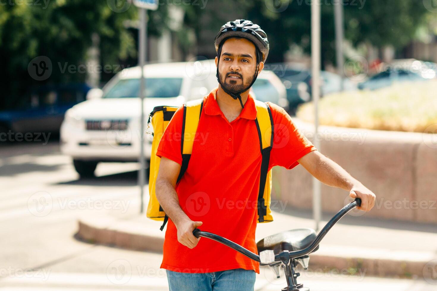 Delivery to home or office. Young man with beard in protective helmet with large yellow backpack with bicycle looking for the address of client, outdoors, free space photo