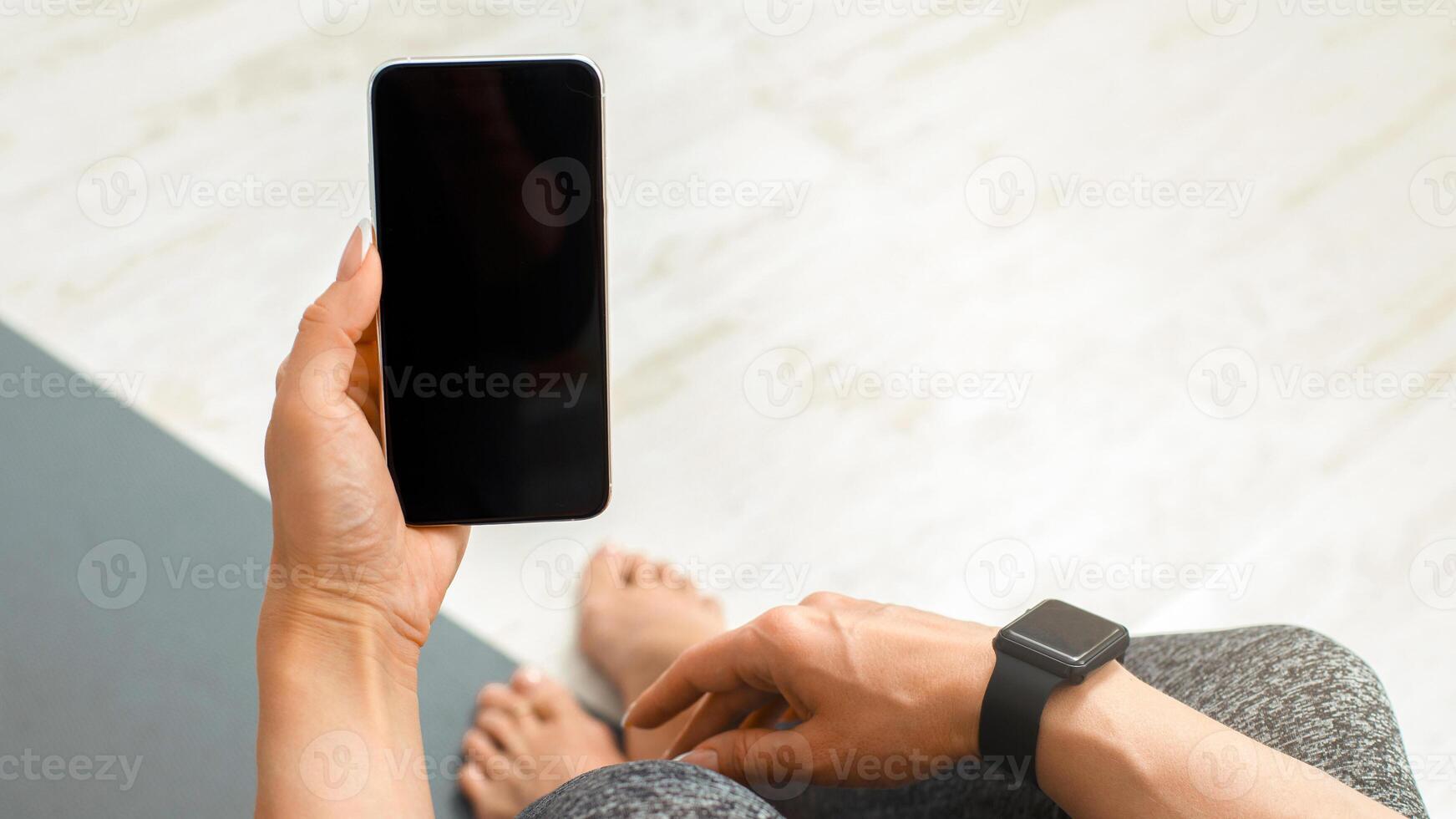 Modern workout at home with gadgets. Woman in sweatpants with smartwatch, holds in hand phone with blank screen, sitting on mat on floor, pov, close up, copy space photo