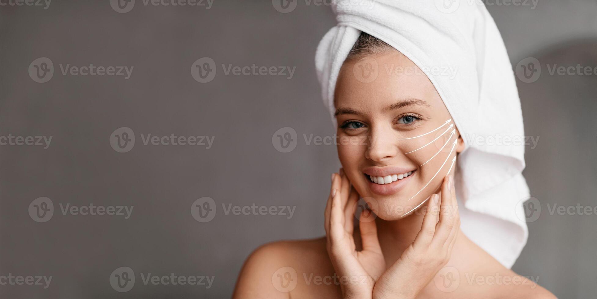 A woman with a towel on her head smiles while pampering her face. She gently massages her skin, embracing self-care and anti-aging practices in a soothing environment, copy space photo