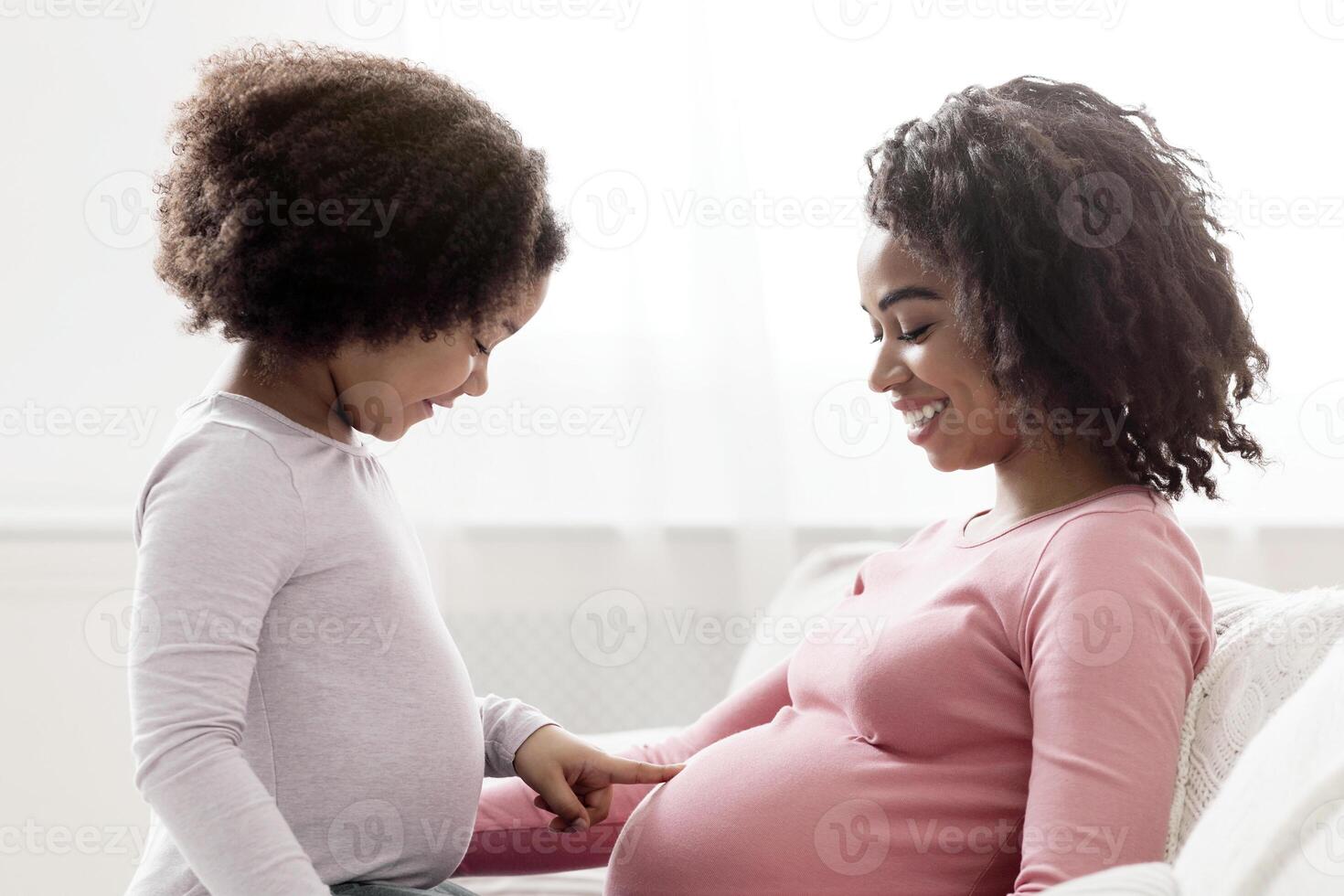 Curious little black girl touching her pregnant mother's belly, sitting on couch at home together, side view with free space photo