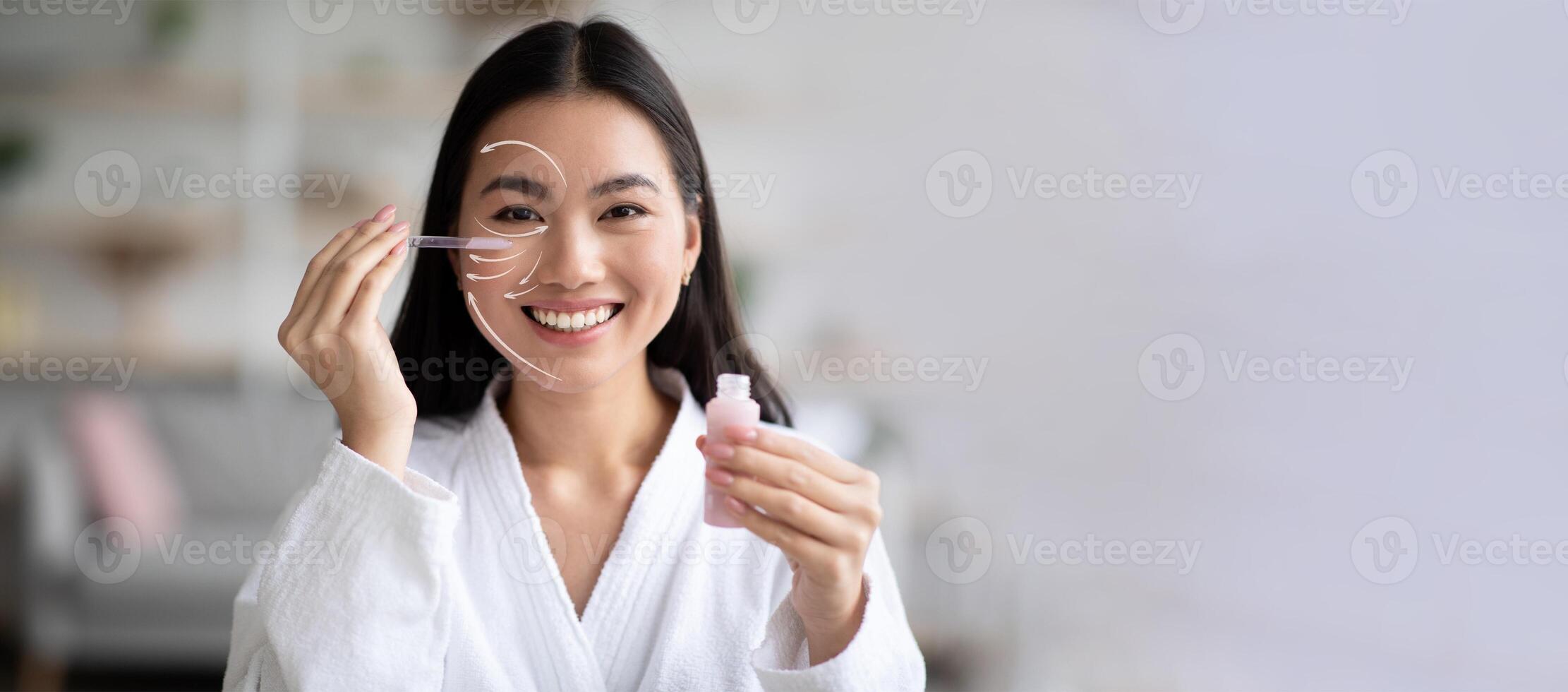 Asian woman is happily applying a serum to her face using a dropper while sitting in a stylish, well-lit room. She enjoys her skincare routine with a focus on rejuvenation and pampering, copy space photo