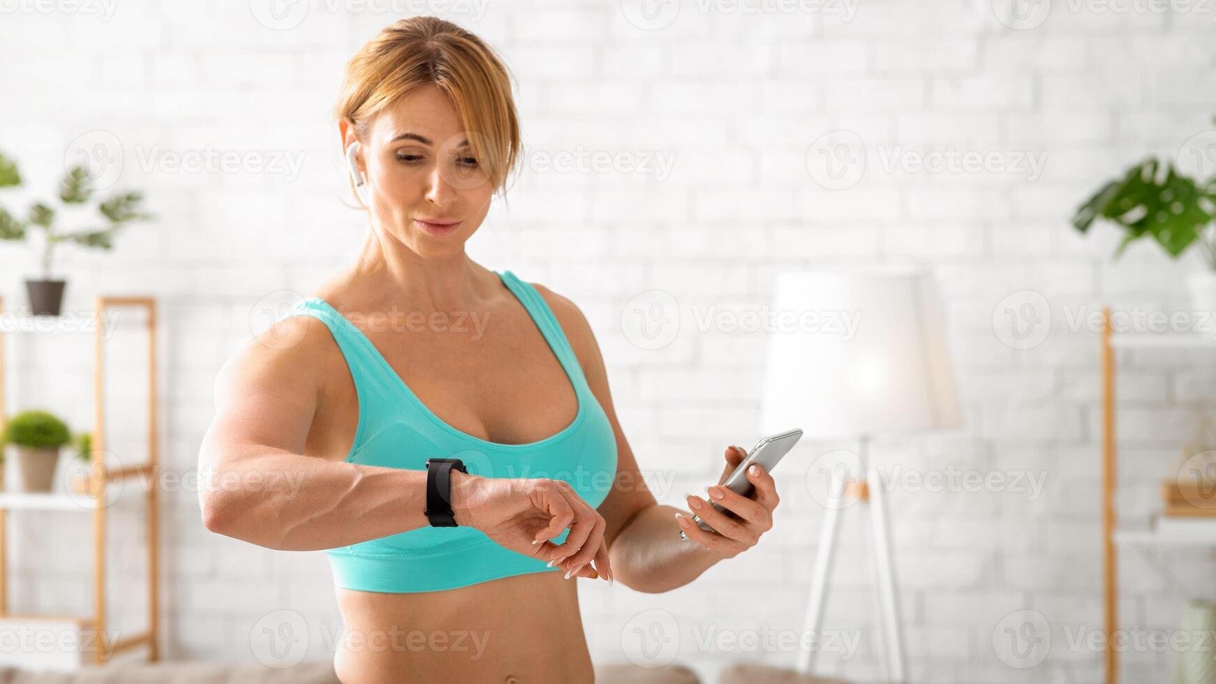 Home fitness workout. Beautiful muscular adult woman in sportswear with smartphone in hand checks fitness tracker in living room interior photo