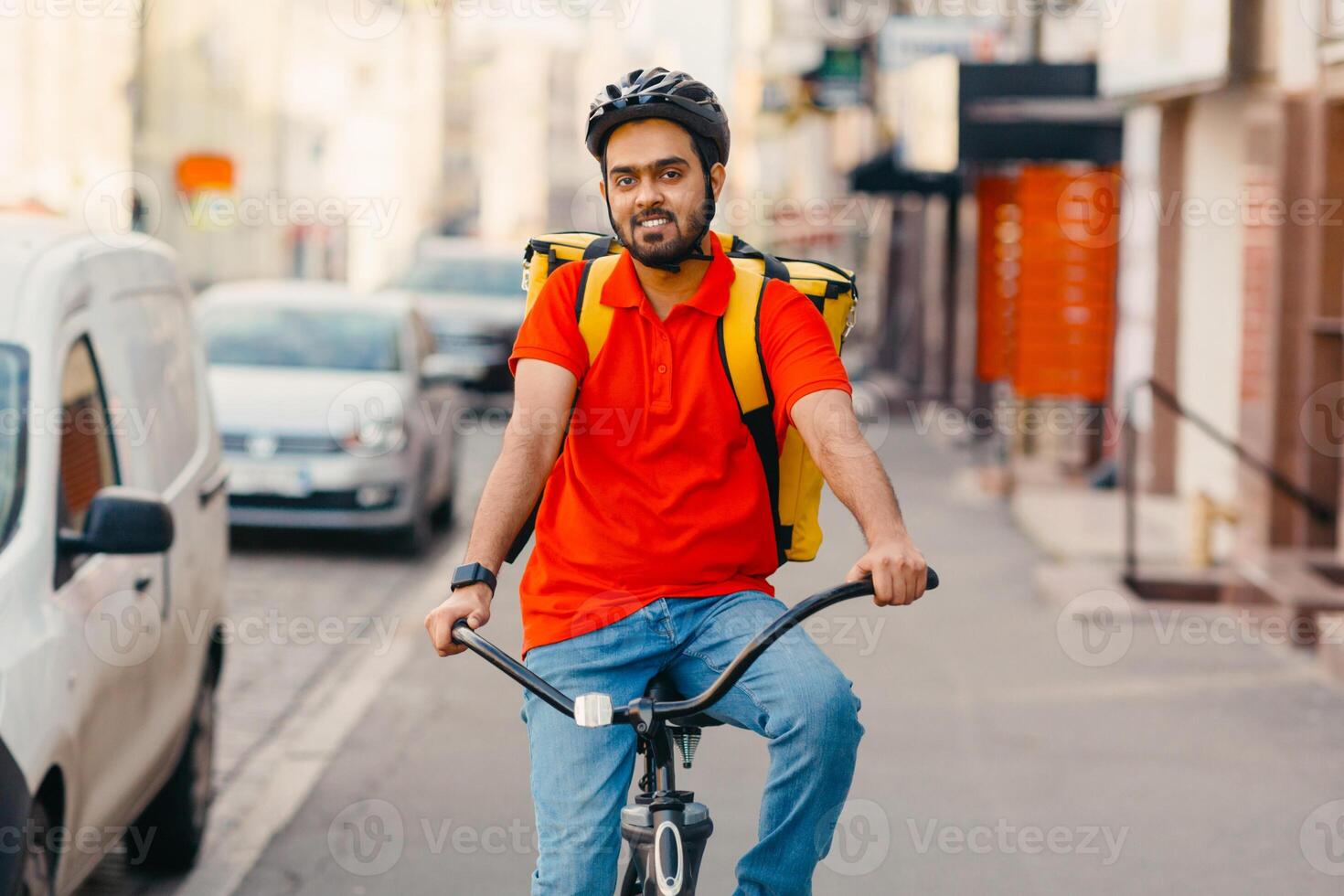 Online delivery service at home. Friendly courier in helmet with yellow backpack on bicycle rides down the street in city, free space photo