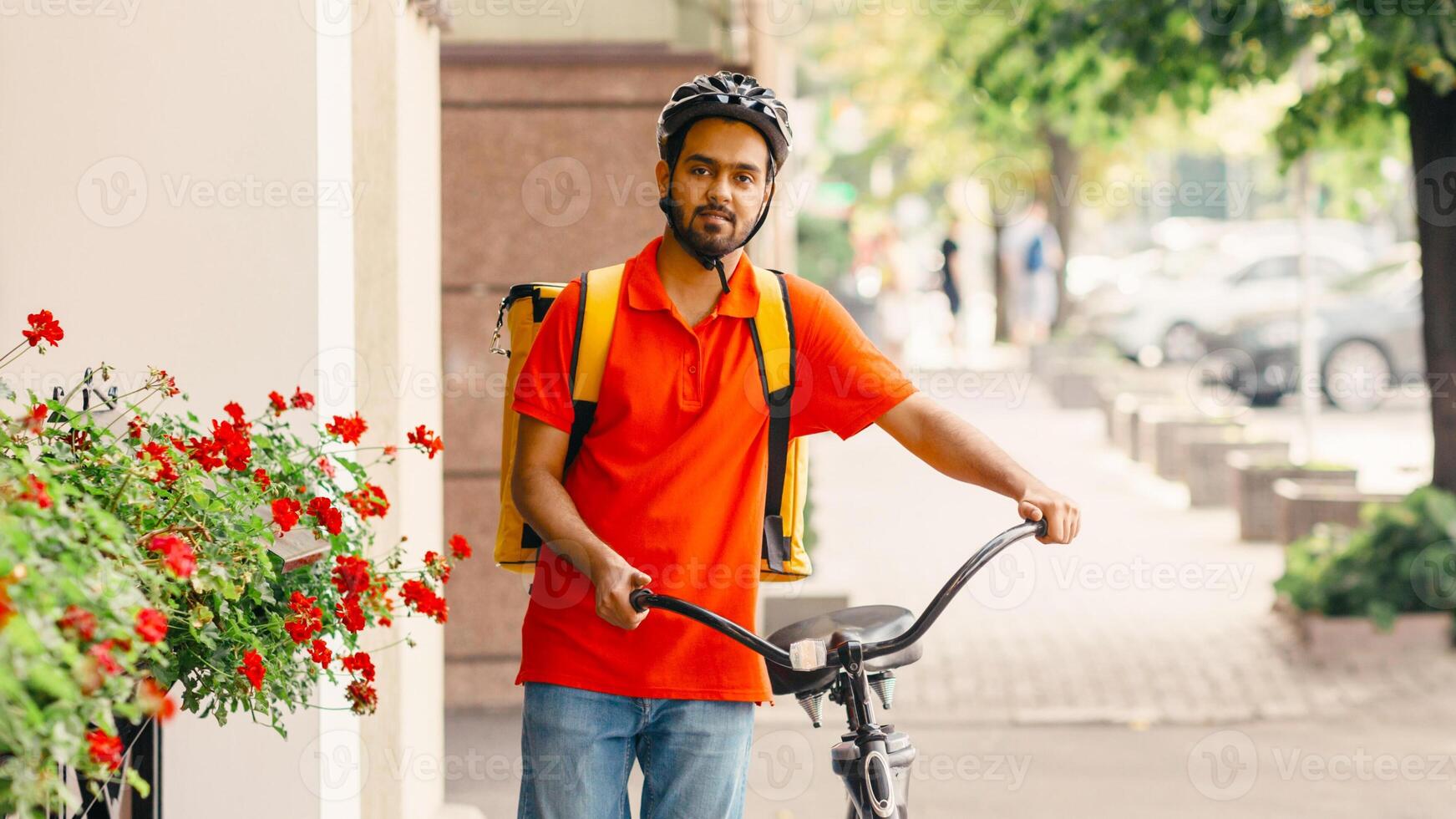 Active physical work in city. Guy curier with beard with yellow backpack in protective helmet, holds bicycle and walks down the street photo
