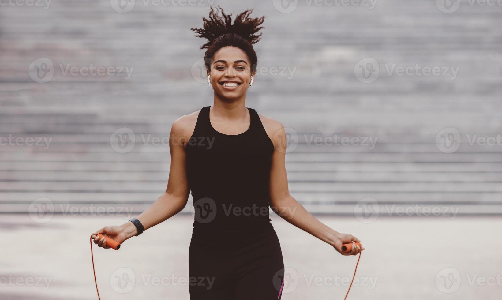 Athlete exercising outside in sunny park. Smiling african american girl in sportswear training with jumping rope on city staircase background, free space photo