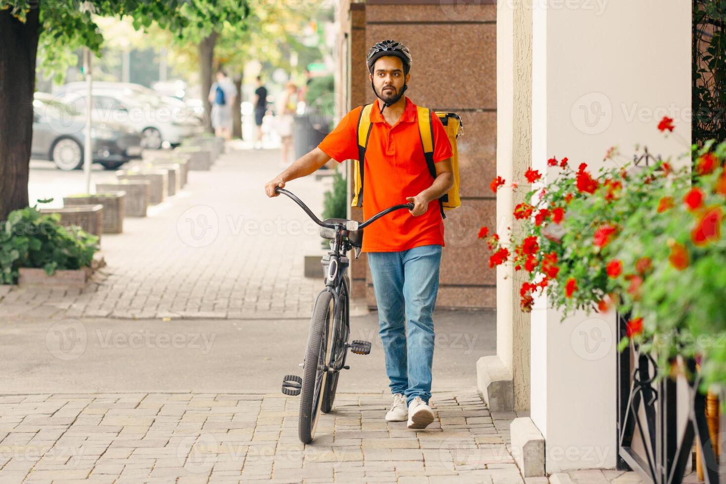 Sports active lifestyle and eco transport in city in summer. Serious young indian male deliveryman in uniform and protective helmet, with big backpack and bicycle walking down the street, outdoor photo