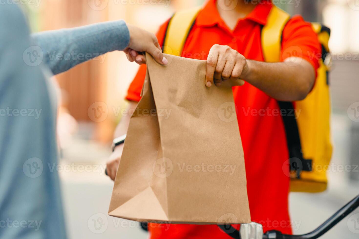 Courier gives fresh lunch to woman, takeaway restaurant food delivery. Young man in orange t-shirt with thermal backpack and bicycle give paper eco bag to client near house, empty space, cropped photo